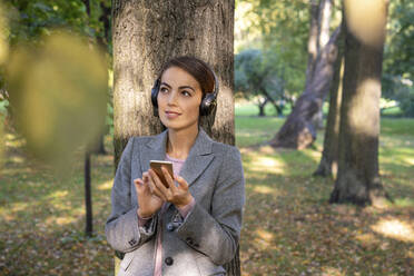 Thoughtful businesswoman listening music while holding smart phone in park during autumn - VPIF03233