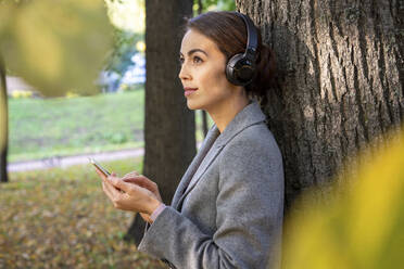 Weibliche Unternehmerin, die im Herbst im Park mit ihrem Smartphone Musik hört und darüber nachdenkt - VPIF03234