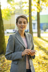 Beautiful businesswoman holding smart phone while standing in park during autumn - VPIF03238