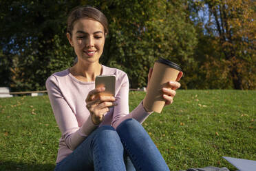 Lächelnde Unternehmerin, die ein Smartphone benutzt, während sie mit einer Kaffeetasse im Herbst im Park sitzt - VPIF03254