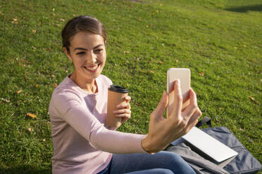 Glückliche Geschäftsfrau, die ein Selfie mit ihrem Smartphone macht, während sie im Herbst auf einer Wiese im Park sitzt - VPIF03255