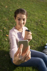 Junge Unternehmerin, die ein Selfie mit ihrem Smartphone macht, während sie im Herbst auf einer Wiese im Park sitzt - VPIF03256