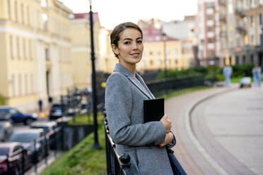 Lächelnde Unternehmerin hält Tagebuch und lehnt sich an ein Geländer in der Stadt - VPIF03262