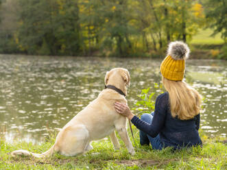 Blond woman sitting with dog at lakeshore during autumn - STSF02683