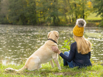 Blonde Frau sitzt mit Hund am Seeufer im Herbst - STSF02683