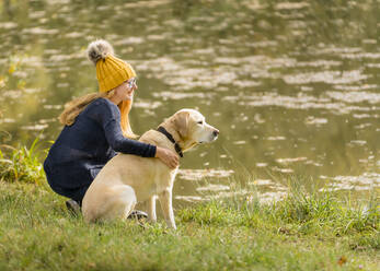 Junge Frau sitzt mit Labrador Retriever am Seeufer im Herbst - STSF02684