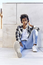 Confident young Afro woman listening music while sitting against retaining wall - VEGF03145