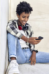 Beautiful young female hipster using smart phone while sitting against retaining wall - VEGF03148