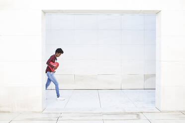 Junge Frau hält roten Ballon in Herzform, während sie gegen eine weiße Wand läuft - VEGF03159