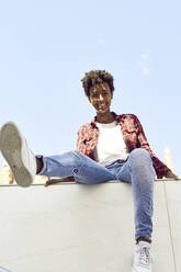 Fröhliche junge Frau sitzt auf einer Stützmauer vor blauem Himmel - VEGF03204