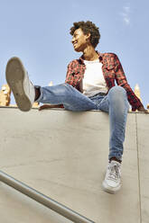 Junge Frau schaut weg, während sie auf einer Stützmauer vor blauem Himmel sitzt - VEGF03205