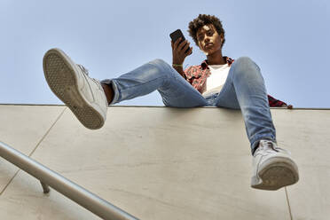 Young female hipster using smart phone while sitting on retaining wall against blue sky - VEGF03207