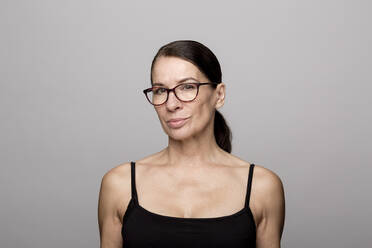 Smiling mature woman in black top wearing eyeglasses against gray background - FLLF00521