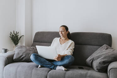 Smiling woman video calling from laptop while sitting on sofa at home - DMGF00315