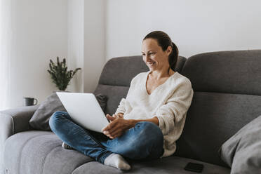 Smiling woman sitting cross-legged using laptop on sofa in living room - DMGF00317