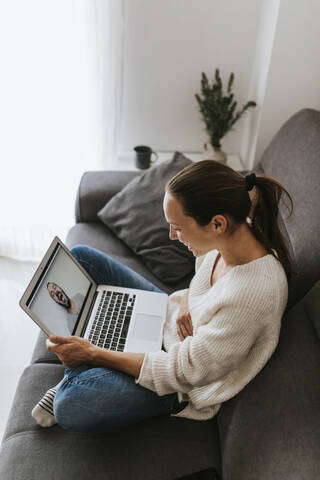 Glückliche Frau sitzt mit der Hand auf dem Bauch und lacht während eines Videoanrufs eines Freundes zu Hause, lizenzfreies Stockfoto