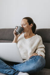 Frau trinkt Kaffee und benutzt einen Laptop zu Hause - DMGF00323