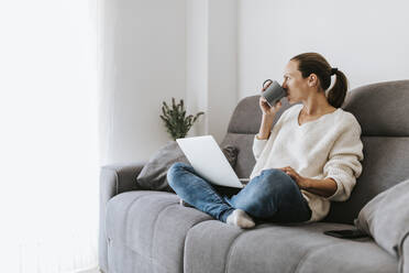 Frau, die wegschaut und Kaffee trinkt, während sie mit ihrem Laptop zu Hause auf dem Sofa sitzt - DMGF00324