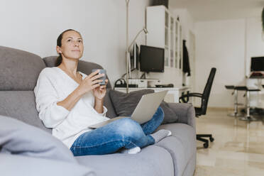 Nachdenkliche Frau, die mit Kaffeetasse und Laptop auf dem Sofa im Wohnzimmer sitzt und wegschaut - DMGF00326