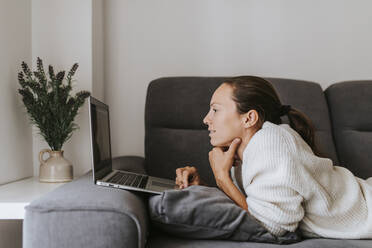 Frau benutzt einen Laptop, während sie auf dem Sofa im Wohnzimmer liegt - DMGF00327