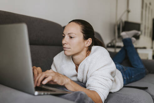 Kaukasische Frau, die einen Laptop benutzt, während sie auf dem Sofa liegt - DMGF00328