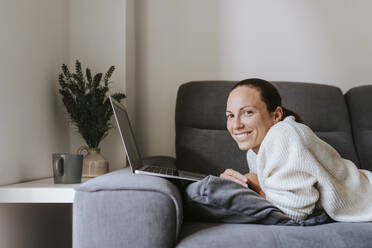 Lächelnde Frau mit Laptop auf dem Sofa im Wohnzimmer liegend - DMGF00330