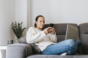 Woman text messaging through smart phone while sitting with laptop on sofa - DMGF00333