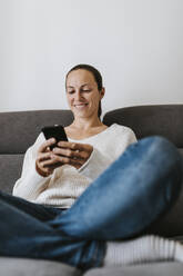 Happy woman using phone on sofa at home - DMGF00334