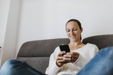 Smiling woman using phone on sofa at home - DMGF00335