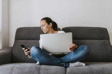 Frau schaut auf ihr Smartphone, während sie mit ihrem Laptop auf dem Sofa sitzt - DMGF00336