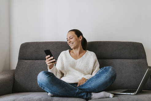 Lächelnde Frau, die ein Smartphone benutzt, während sie mit einem Laptop auf dem Sofa sitzt - DMGF00337