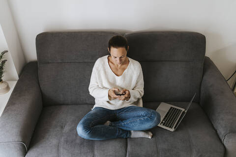 Frau, die ein Mobiltelefon benutzt, während sie mit einem Laptop auf dem Sofa sitzt, lizenzfreies Stockfoto