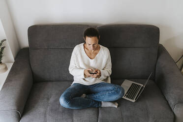 Frau, die ein Mobiltelefon benutzt, während sie mit einem Laptop auf dem Sofa sitzt - DMGF00338