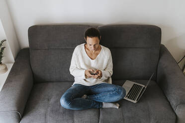 Woman using mobile phone while sitting with laptop on sofa - DMGF00338
