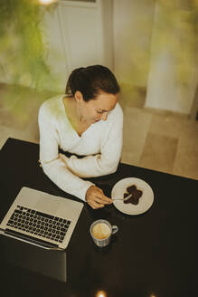 Frau isst Schokoladen-Brownie, während sie mit Laptop und Kaffee an der Kücheninsel sitzt - DMGF00345