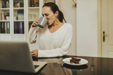 Frau trinkt Kaffee und schaut auf einen Laptop in einer beleuchteten Küche - DMGF00349