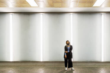 Young woman with cool attitude standing against illuminated wall in tunnel during pandemic - IFRF00097