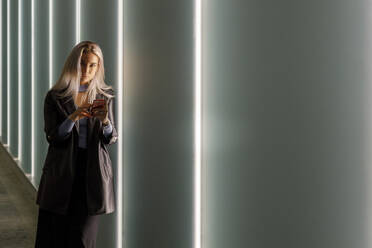 Beautiful young woman holding smart while leaning on illuminated wall in tunnel - IFRF00102