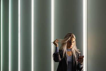 Attractive woman holding hair in hand with smart phone while standing against illuminated wall - IFRF00104