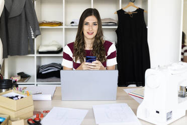 Young businesswoman holding smart phone while sitting with laptop at studio - GIOF09748