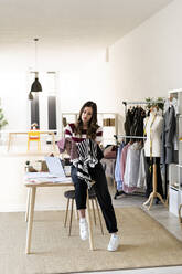 Young female design professional examining tops while sitting on desk at studio - GIOF09759