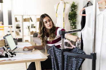 Young businesswoman with laptop looking at tops arranged on rack in studio - GIOF09765