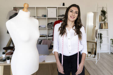 Young tailor with measure tape around her neck standing by mannequin at studio - GIOF09769