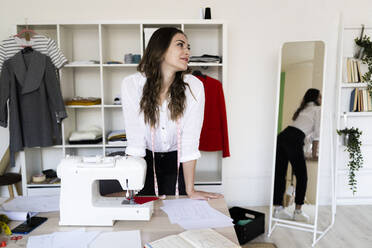 Female fashion designer looking away while leaning on desk in studio - GIOF09772