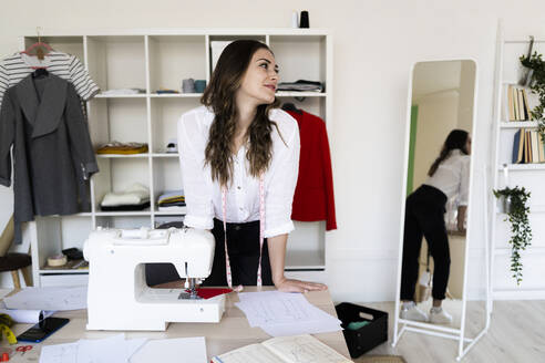Weibliche Modedesignerin schaut weg, während sie sich im Atelier auf den Schreibtisch stützt - GIOF09772