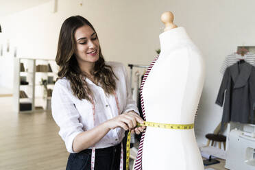 Smiling female designer measuring waist of mannequin while standing at studio - GIOF09776