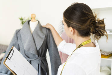 Weibliche Designerin, die ein Klemmbrett hält, während sie einen Wintermantel im Studio analysiert - GIOF09778