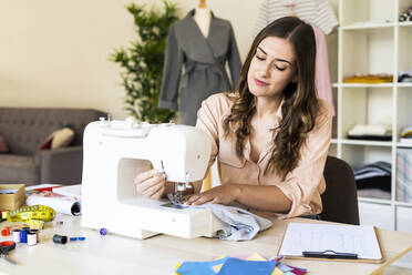 Smiling young female creative professional using sewing machine while sitting at studio - GIOF09793