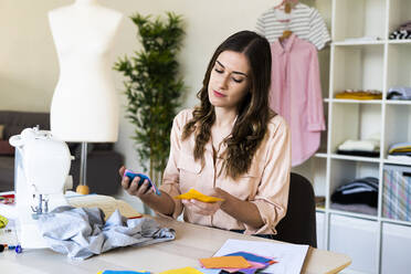 Beautiful female designer examining fabric swatches while sitting in studio - GIOF09799