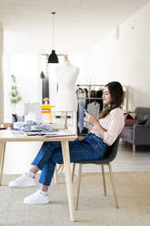 Junge Designerin benutzt ein Smartphone, während sie auf einem Stuhl im Studio sitzt - GIOF09805